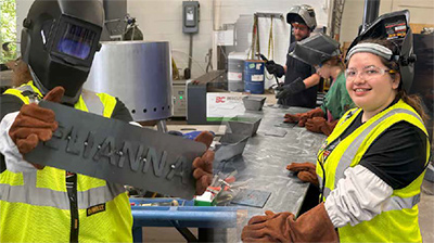 Workers in safety gear holding a sheet metal sign in a workshop.