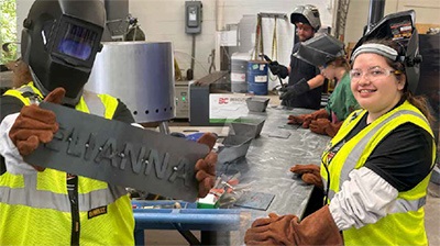 Workers in safety gear holding a sheet metal sign in a workshop.